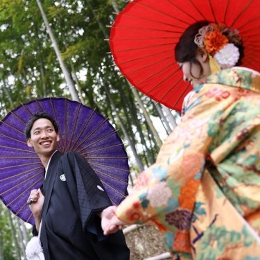 竹林で紫と赤の和傘を差して手をつなぐ紋付袴の新郎と色打掛の花嫁。白鳥庭園で撮る春の和装前撮り