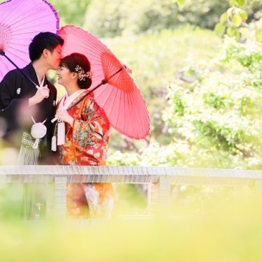池の木の橋の上で赤い和傘の下で花嫁の頬にキスする紋付袴の新郎と色打掛の花嫁。白鳥庭園の和装前撮り