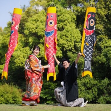 こいのぼりの下でひざまずいて手を伸ばす紋付袴の新郎と笑う色打掛の花嫁。白鳥庭園の初夏の和装前撮り