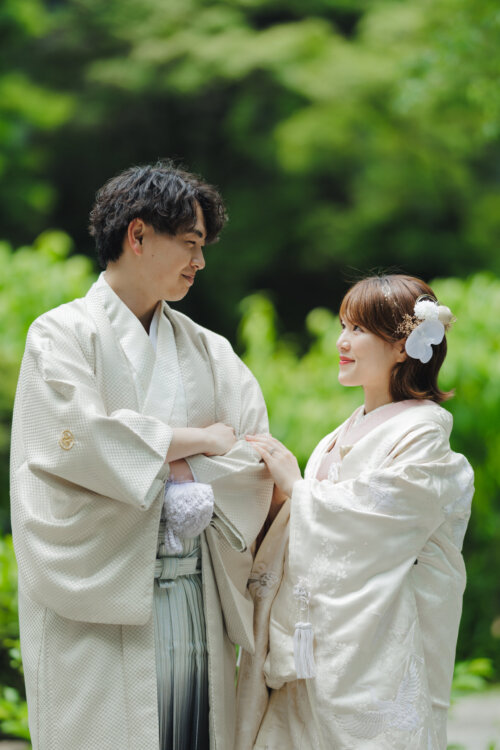 緑の木立を背に見つめ合う、白紋付袴の新郎とピンクの掛下を合わせた白無垢の花嫁の和装前撮り