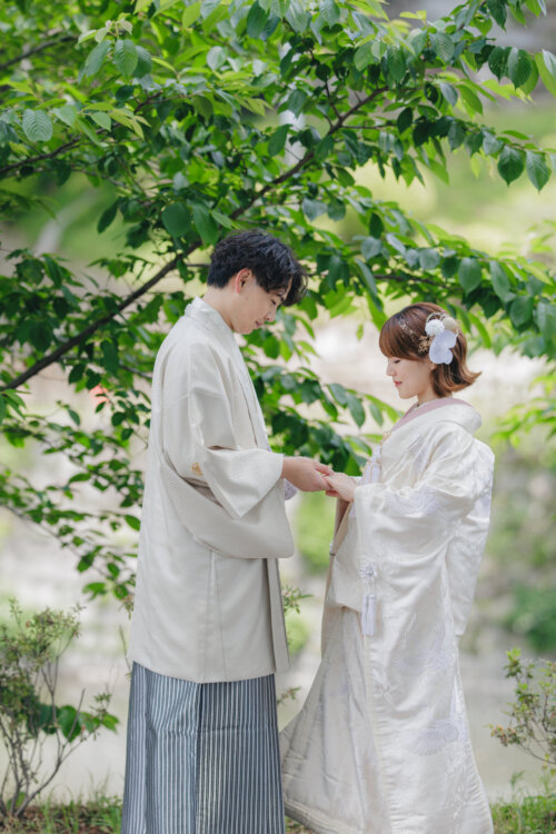 新緑の枝の下で両手を取り合う、白紋付袴の新郎と白無垢の花嫁。初夏の公園でのフォトウェディング