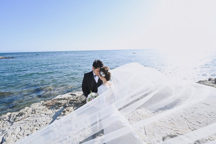 海が見える砂浜でベールが綺麗に風に吹かれる新郎新婦