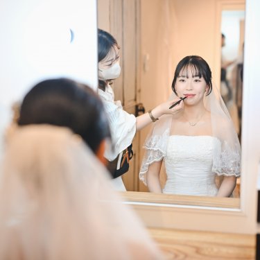 鏡の前でベール姿の花嫁がリップを仕上げてもらうヘアメイクの様子。フォトウェディング当日の支度
