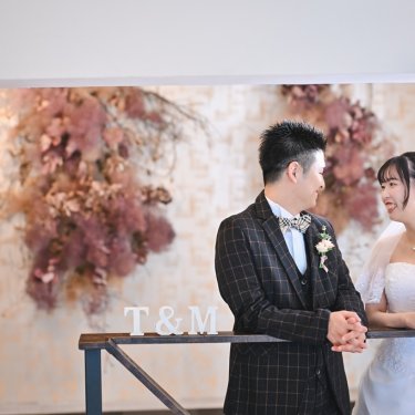 ドライフラワーの壁の前で手すりにもたれ見つめ合う新郎新婦。アトリエスタジオのウェディングフォト