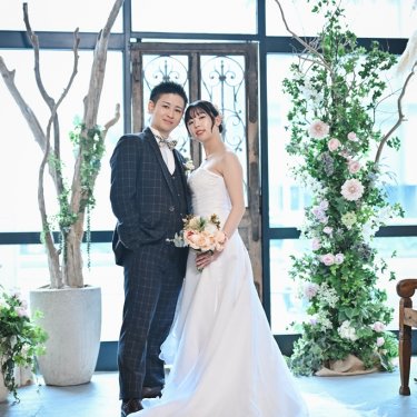 大きな窓とグリーンに囲まれたアトリエスタジオで寄り添う新郎新婦。ロングトレーンのウェディングドレスの前撮り