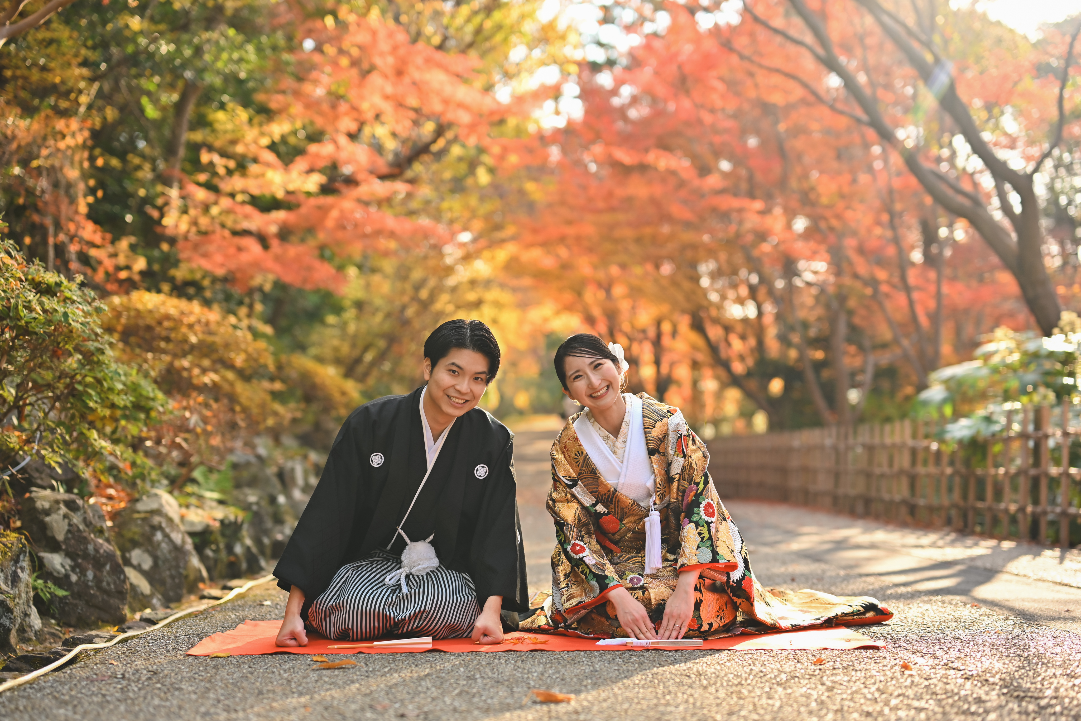 紅葉の並木道で緋毛氈に座って笑う、紋付袴の新郎と色打掛の花嫁。東山植物園で撮る秋の和装前撮り