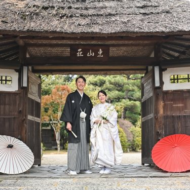 東山荘の茅葺の門の下に並ぶ新郎新婦と、左右に置いた白と赤の和傘。シンメトリーな構図の和装ロケーションフォト