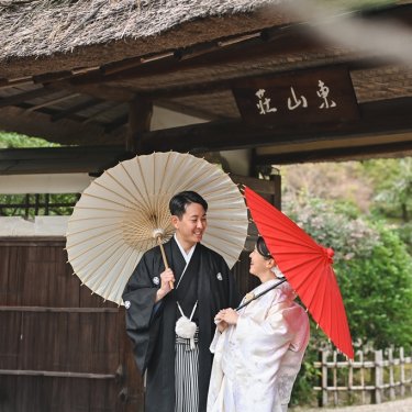 東山荘の茅葺の門の前で、白と赤の和傘を差して見つめ合う紋付袴の新郎と白無垢の花嫁。和装前撮りの定番ポーズ