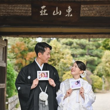 '東山荘'の額がある門の下で、赤ちゃんの頃の写真を手に笑う紋付袴の新郎と白無垢の花嫁。思い出を形にする和装前撮り