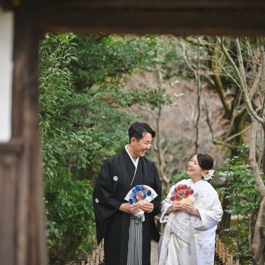 門の向こうの緑の中で、扇子を手に見つめ合う紋付袴の新郎と白無垢の花嫁。和装ロケーションフォトの一枚