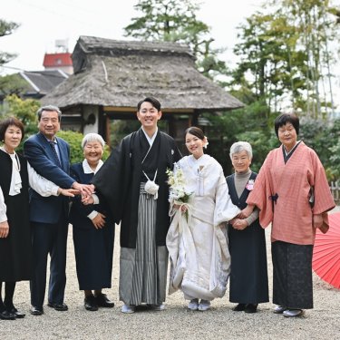 茅葺の門の前で、和傘を置いて手をつなぐ新郎新婦とご家族。東山荘の和装前撮りで残す三世代の家族写真