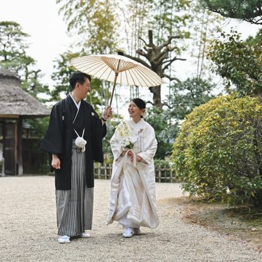 名古屋の東山荘の茅葺の門を背に、和傘を差して歩く紋付袴の新郎と白無垢の花嫁。砂利敷きの庭での和装前撮り