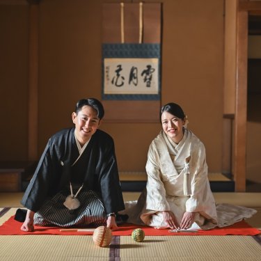 緋毛氈を敷いた和室で、掛け軸の前に正座する紋付袴の新郎と白無垢の花嫁。手まりを添えた和装前撮り