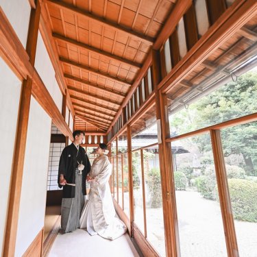 日本庭園に面した長い廊下に立つ白無垢の花嫁と紋付袴の新郎。東山荘の木造建築を生かした引きの和装前撮り