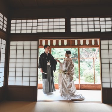 東山荘の障子の和室の奥、庭へ開いた戸口に並ぶ紋付袴の新郎と白無垢の花嫁。奥行きのある構図の和装前撮り