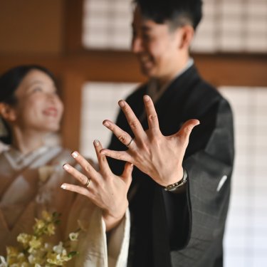 指輪をつけた手を重ねて見せ合い、笑い合う紋付袴の新郎と白無垢の花嫁。和室の障子をぼかした和装前撮り