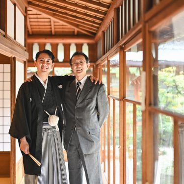 ガラス戸の続く廊下で、肩を組んで笑う紋付袴の新郎とお父様。家族写真も残せる和装前撮りの一枚