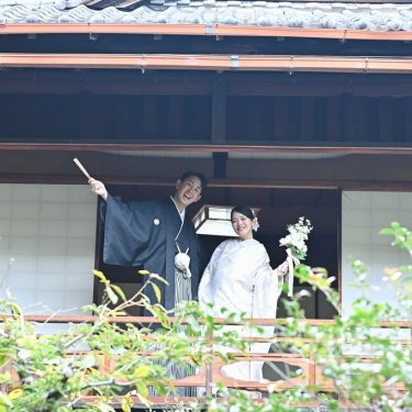 緑の木々越しに見える縁側で、扇子とブーケを掲げて笑う新郎新婦。古い日本家屋で撮る和装ロケーションフォト