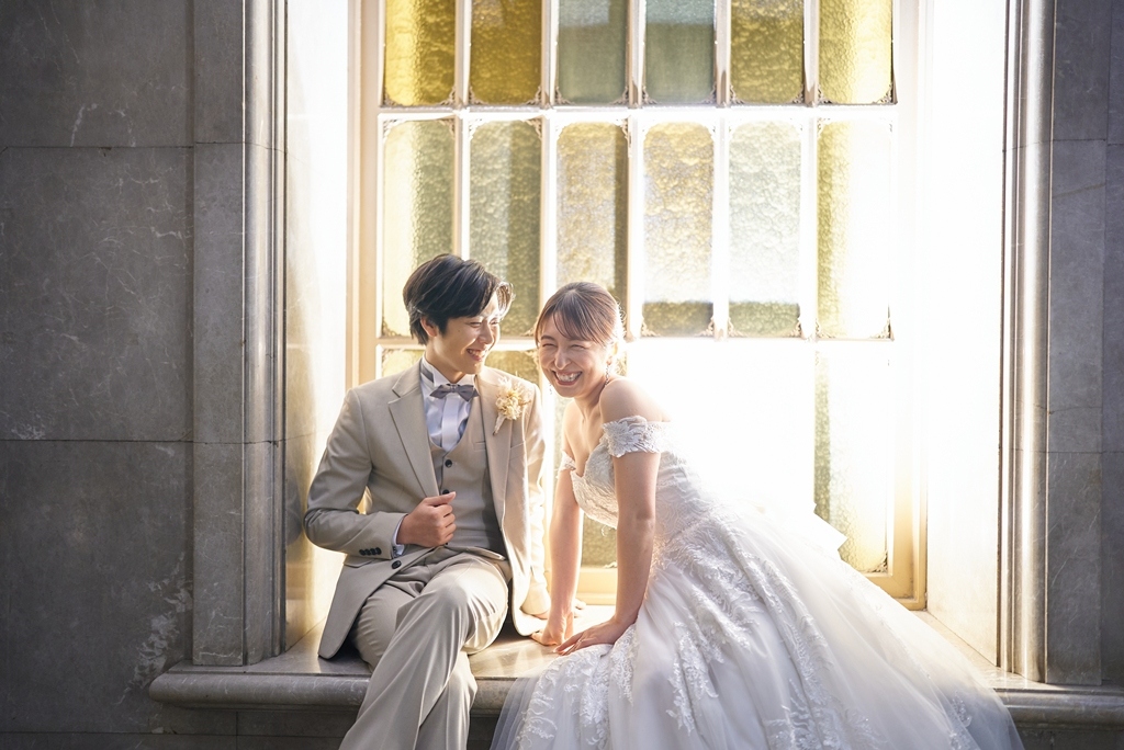 ステンドグラス風の窓辺に腰掛けて笑い合うベージュスーツの新郎とオフショルダードレスの新婦