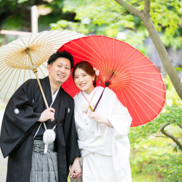 赤と白の和傘を重ねるように差し、カメラに笑顔を向ける白無垢の花嫁と紋付袴の新郎。緑が映える和装前撮り