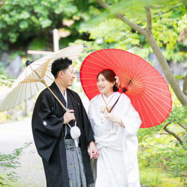 木漏れ日の庭で赤と白の和傘を差し、手をつないで見つめ合う新郎新婦。宗節庵で撮った白無垢の前撮り