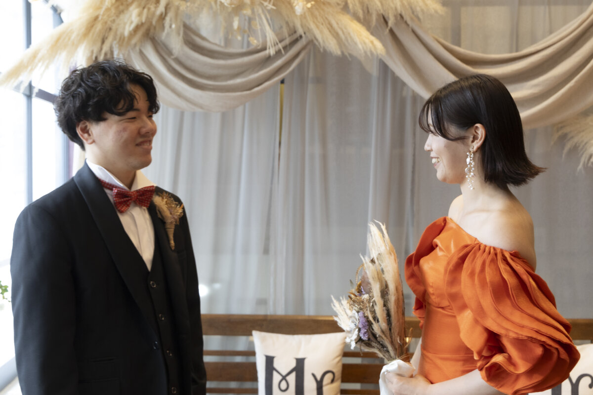 パンパスグラスのドレープの前で振り返った新郎とオレンジのカラードレスの花嫁が見つめ合うファーストミートのスタジオフォト