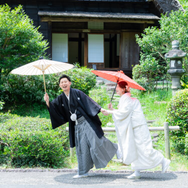 石灯籠のある庭で、和傘を差した白無垢の花嫁が紋付袴の新郎の背中を押す遊び心あるワンシーン。宗節庵の和装前撮り