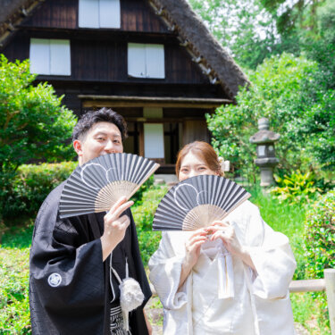 茅葺き屋根の古民家の前で、扇子で口元を隠してほほえむ新郎新婦。小物を使った和装前撮りのポーズ