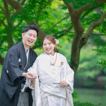 池のほとりの新緑の下で、顔を寄せて笑う紋付袴の新郎と白無垢の花嫁。初夏の和装前撮り