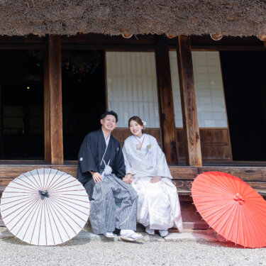 茅葺き屋根の古民家の正面で、両脇に和傘を置いて縁側に座る新郎新婦。古民家の佇まいを生かした和装の結婚写真