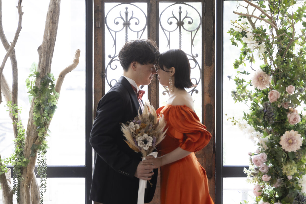 アイアンの窓と花のアーチの前で額を寄せ合う新郎とオレンジのカラードレスの花嫁。名古屋のスタジオで撮るウェディングフォト