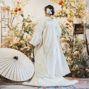 ドライフラワーの装飾の前で白い和傘を置き、振り返る白無垢の花嫁の後ろ姿。スタジオで撮る和装前撮り