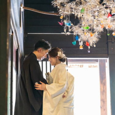 折り鶴が吊るされた古民家の軒下で、おでこを寄せ合う紋付袴の新郎と白無垢の花嫁。逆光がやわらかい和装フォトウェディング