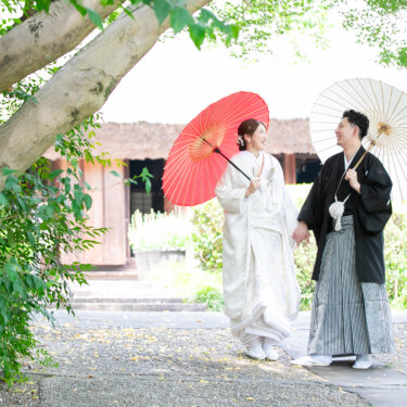 木々の枝越しに、赤と白の和傘を差して手をつなぎ歩く新郎新婦を写した一枚。宗節庵の庭で撮る白無垢の前撮り