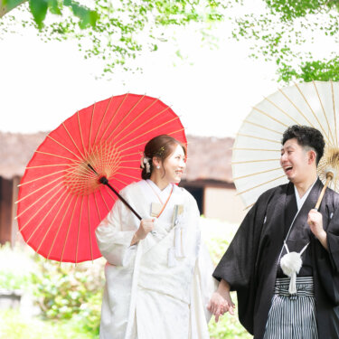 茅葺き屋根の古民家を背に、和傘を手に顔を見合わせて歩く白無垢の花嫁と紋付袴の新郎。緑あふれる和装ロケーションフォト