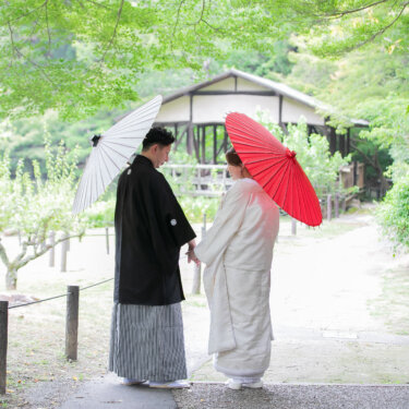 茅葺きの東屋へ続く小道で、和傘を差して手をつなぐ紋付袴の新郎と白無垢の花嫁の後ろ姿。緑に包まれた和装の結婚写真