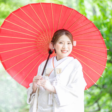 赤い和傘を差して笑顔を見せる白無垢の花嫁のアップ。新緑の中で撮った宗節庵の和装前撮り