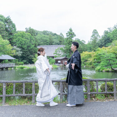 池と茅葺き屋根の建物を背景に、柵の前で向かい合う白無垢の花嫁と紋付袴の新郎。開放的な庭園で撮る和装前撮り
