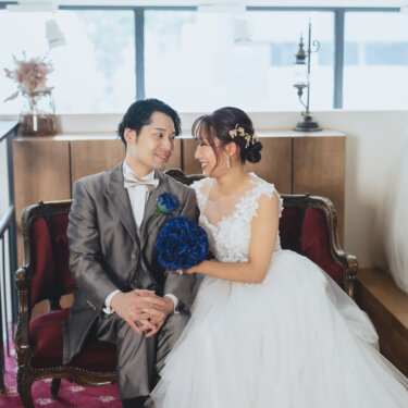 赤いソファに並んで座り、見つめ合って笑うグレーのタキシードの新郎とレースのウェディングドレスの花嫁の前撮り
