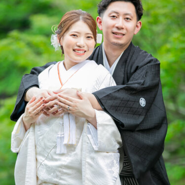 新緑を背に、新郎が後ろから白無垢の花嫁を包み込むバックハグのポーズ。指輪が光る和装前撮りのアップ