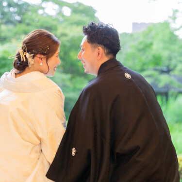 庭の緑を眺めながら顔を寄せ合う白無垢の花嫁と紋付袴の新郎の後ろ姿。和装前撮りで人気の背中越しのカット