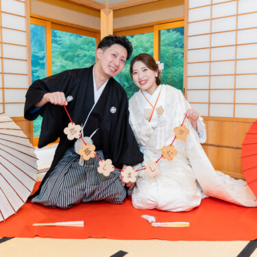 赤い毛氈の上に並んで座り、梅の形のガーランドを掲げてほほえむ新郎新婦。和傘を両脇に置いた和室の前撮り