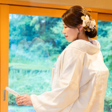 緑が見える大きな窓辺で、扇子を手にうつむく白無垢の花嫁の横顔。髪飾りを添えたまとめ髪が映える和装前撮り