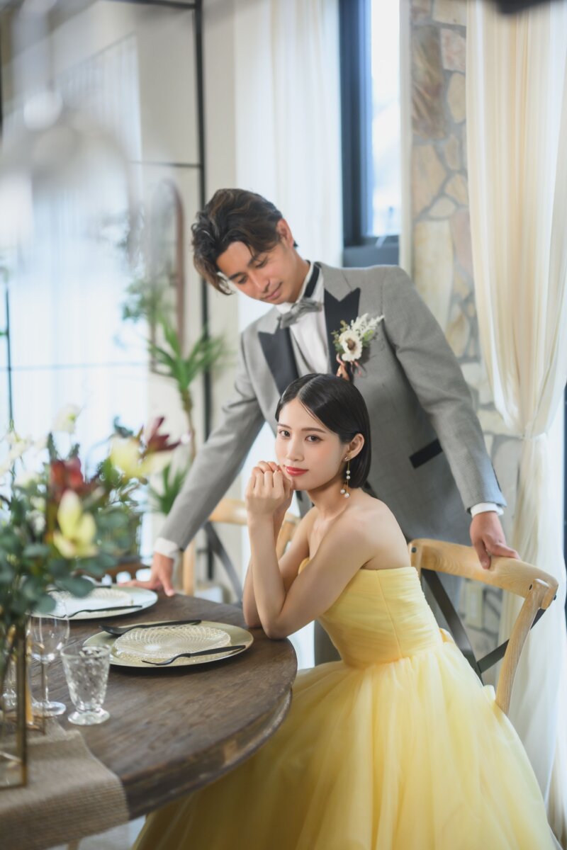 ダイニングテーブルで頬杖をつくイエローのカラードレスの花嫁と、後ろから寄り添うグレーのタキシードの新郎。名古屋のスタジオフォト