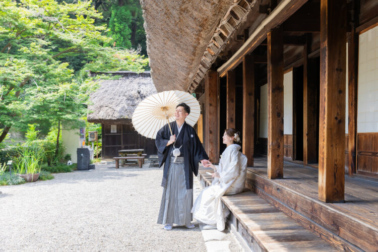 茅葺き屋根の古民家の長い縁側で、座る白無垢の花嫁に和傘を差した新郎が話しかける一枚。宗節庵の和装フォトウェディング