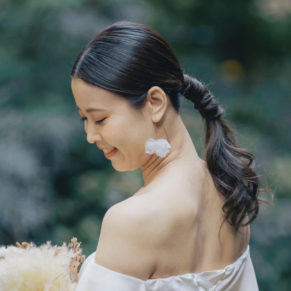 センターパートでまとめたポニーテールと、花モチーフの揺れるイヤリング。オフショルダードレス姿の花嫁が微笑む横顔クローズアップ。