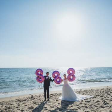 青空の下の砂浜で、ピンクの数字バルーンを持って並ぶ新郎新婦を遠景で写した知多半島の海のウェディングフォト