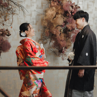 ドライフラワーの壁の前で手すり越しに向き合い、照れ笑いする赤い色打掛の花嫁と紋付袴の新郎。アトリエスタジオで撮る和装前撮り