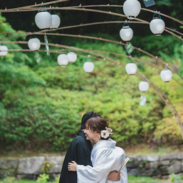 白い提灯のアーチの下で、新郎に抱きついて笑う白無垢の花嫁。夏の庭園で撮る、ハグのポーズが自然な結婚写真