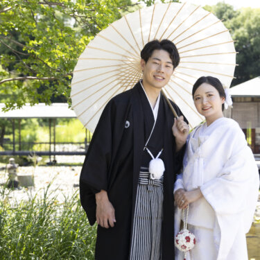 白い番傘をさした紋付袴の新郎と、寄り添う白無垢の花嫁。池と茶屋が見える白鳥庭園で撮る和装フォトウェディング